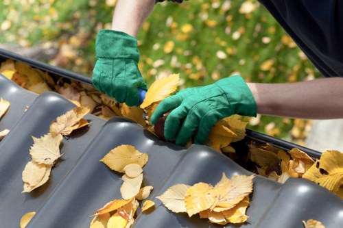 A person wearing green gardening gloves is collecting fallen yellow and brown leaves and placing them into a grey plastic leaf gutter guard. The gutter guard is installed on a sloped roof, which is partially visible in the foreground. In the background, there is a garden area with green foliage and some blurred yellow leaves. The scene suggests maintenance or clearing work related to outdoor rubbish removal. This type of activity aligns with professional waste removal services offered by businesses like Business Waste Removal Abbey Wood, serving local residents and commercial clients in the Abbey Wood area near postcode SE2. The natural lighting highlights the textures of the leaves and the reflective surface of the gutter guard, providing a clear, detailed view suitable for accessibility and search engine optimization regarding rubbish and waste disposal services in the Kent Thameside region.
