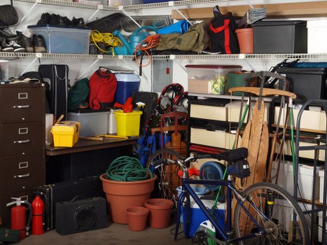 An indoor storage area or garage with a densely packed arrangement of miscellaneous household and outdoor items. In the foreground, there are several large terracotta plant pots of various sizes stacked on the concrete floor, along with a coiled garden hose in a green color. To the right, a blue bicycle with a black saddle and red accents leans against a shelving unit. The shelves above contain a diverse collection of storage boxes, toolkits, and bags, including a large black plastic storage container, a red bag, and a grey bin, all filled with assorted household items. A yellow plastic box and a small red toolbox are also visible on the lower shelf. To the left, a dark metal filing cabinet with multiple drawers stands beside a black rectangular radio or speaker. Various ropes, cords, and garden tools are haphazardly arranged on and hanging from the metal shelves. The environment appears to be a typical cluttered workspace or storage area used for various domestic and gardening purposes, situated in a residential area near Abbey Wood, with a functional, utilitarian atmosphere suitable for waste or rubbish collection planning, provided by Business Waste Removal Abbey Wood.